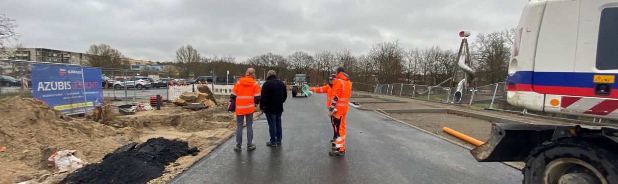 Straßenansicht mit Bauarbeitern in Warnwesten und Baufahrzeug.