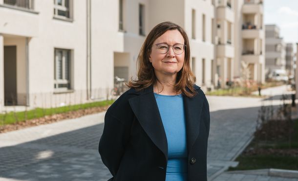 Sandra Jacob Porträt einer Frau mit braunen, halblangen Haaren, Brille, blauem Oberteil und schwarzem Sacko vor einer sonnigen Häuserzeile stehend