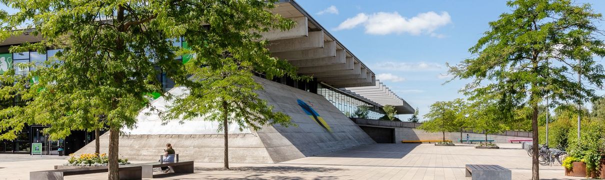 pyramidenartig gebautes Gebäude mit großem Flachdach, vor dem grüne Bäume stehen