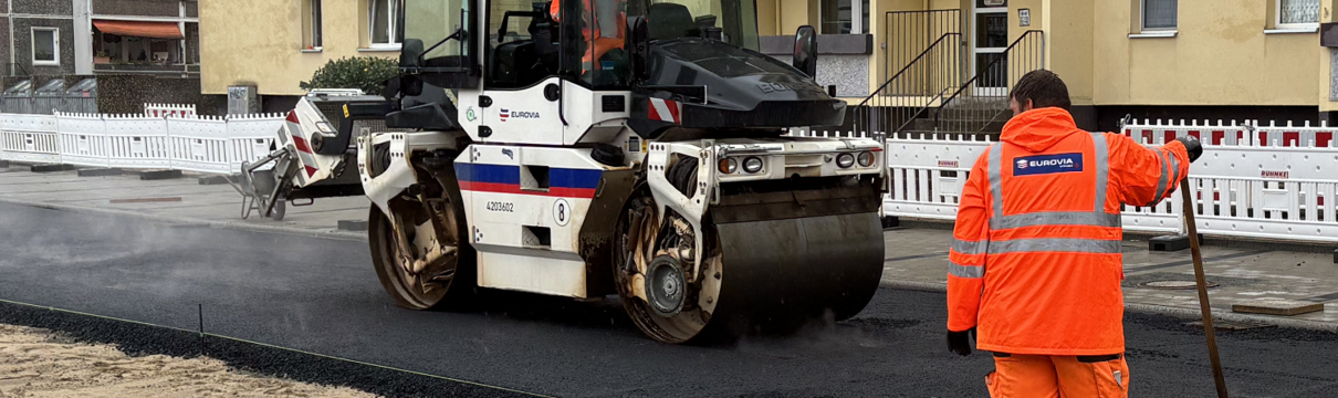 Orange gekleideter Bauarbeiter und ein Baufahrzeug an einer Straße.