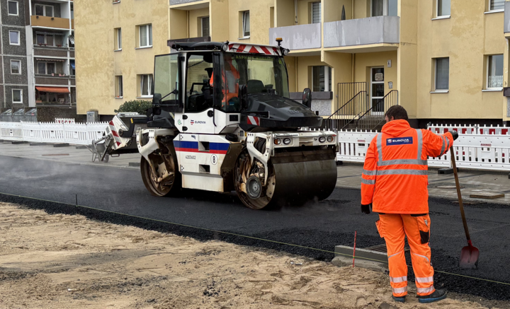 Orange gekleideter Bauarbeiter und ein Baufahrzeug an einer Straße.