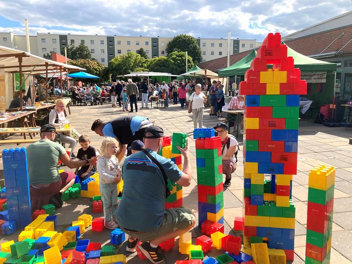 viele bunte große Bausteine werden von Familien verbaut auf einem Platz mit vielen kleinen Marktständen vor einem großen Plattenbau