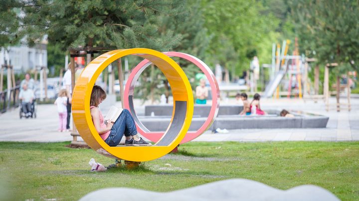 Im Vordergrund sitzt eine Frau in einem großen, gelben Sitzring, dahinter steht ein rosa farbener Ring und es schließt sich ein Spielplatz an