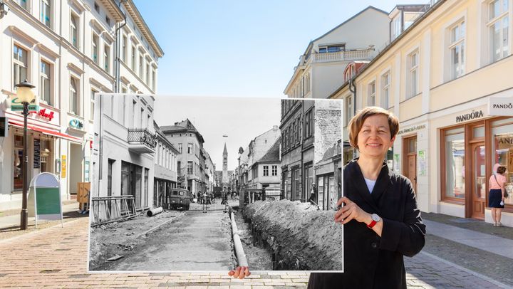 Eine Frau steht auf einer Einkaufsstraße und hält ein großes Schwarz-Weiß-Bild hoch, wie diese vor der Sanierung aussah