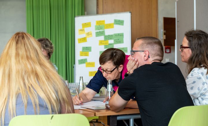 Wand mit vielen, bunten, beschriebenen Klebezetteln, vor der Menschen am Tisch sitzen und zusammenarbeiten