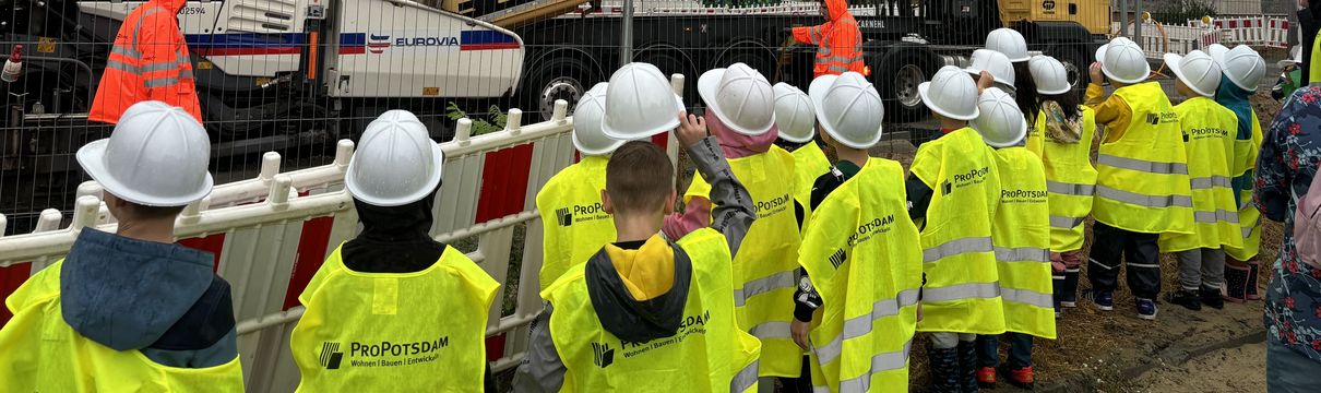 Kinder mit gelber Warnweste und weißen Bauhelmen an einer Baustelle.