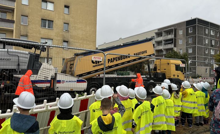 Kinder mit gelber Warnweste und weißen Bauhelmen an einer Baustelle.