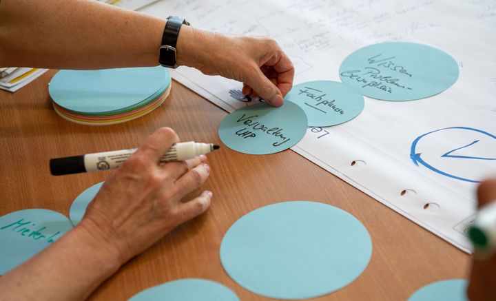 blaue, runde Zettel auf einem Tisch, die gerade von einer Hand beschrieben werden