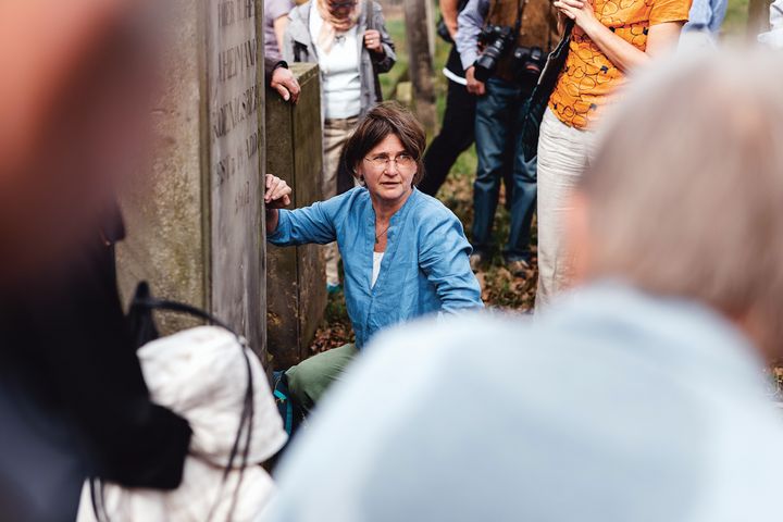 Eine Frau hockt vor einem Grabstein. Um sie herum steht eine Gruppe von Menschen.