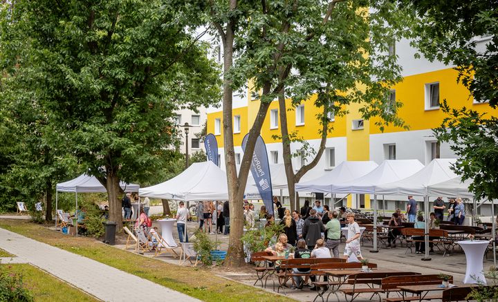 Festliche, weiße Pavillons und hölzerne Sitzgelegenheiten mit vielen Menschen vor einem Plattenbau mit gelben Streifen