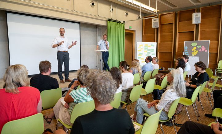 größere Gruppe, die auf grüne Stühlen sitzt hört Mann zu, der auf Podest vor einer Beamerwand steht