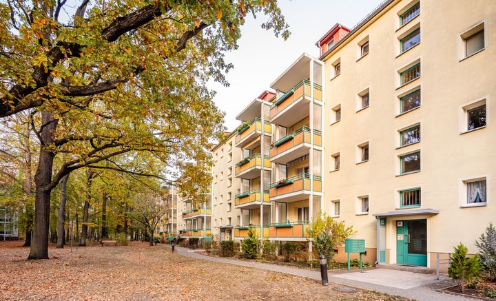 herbstliche Bäume, an denen sich ein Weg entlang eines Plattenbaus mit gelben und orangen Balkonen entlang schlängelt