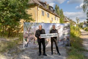 Zwei Männer stehen mit einem Straßenbild vor einem Haus und einer Tafel mit einem Bild.