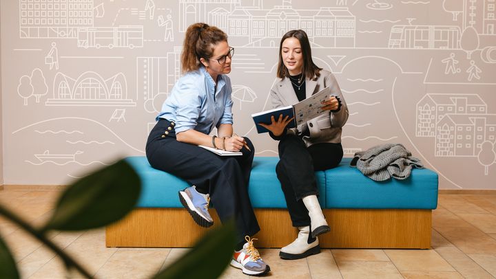 Zwei Frau sitzen auf einer blau bepolsterten Sitzgruppe und eine der beiden zeigt der anderen etwas in einem Booklet