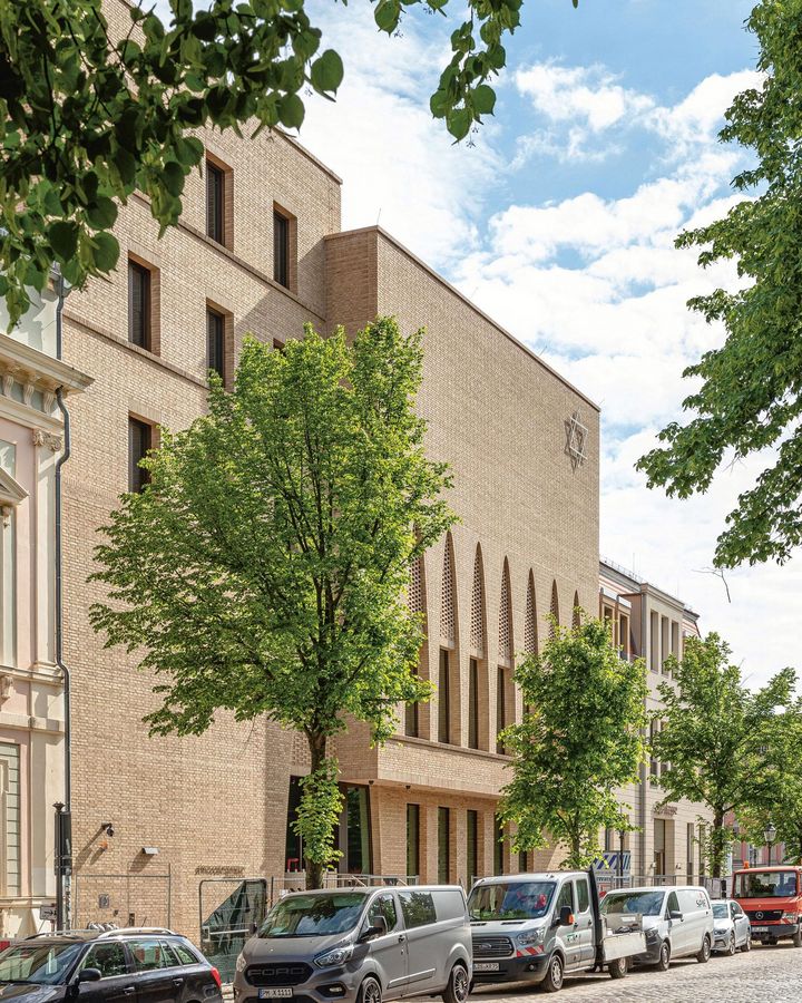 Gebäude entlang einer Straßen mit Kopfsteinpflaster. Davor stehen Häuser und grün-blühende Bäume.