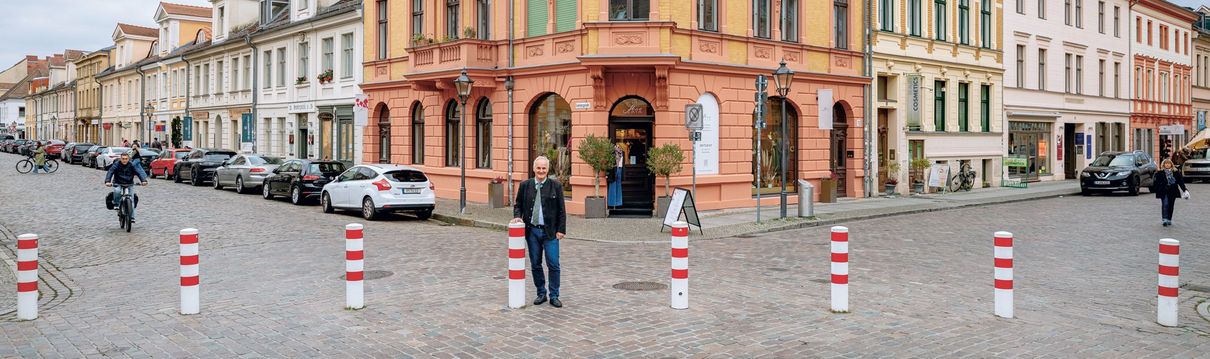 Ein Mann steht an einer Kreuzung. Auf der Straße sind rot-weiß-gestreifte Poller, die den Autoverkehr einschränken. 