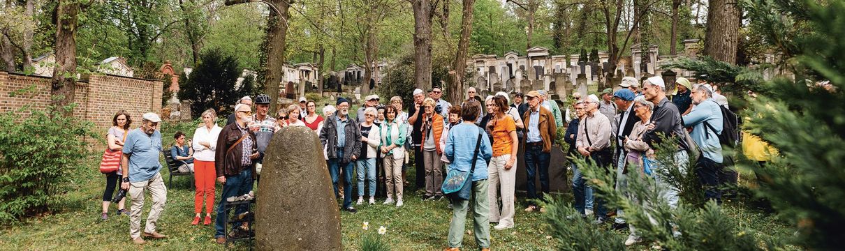 Eine große Menschengruppe steht auf einem Friedhof mit Grabsteinen. 